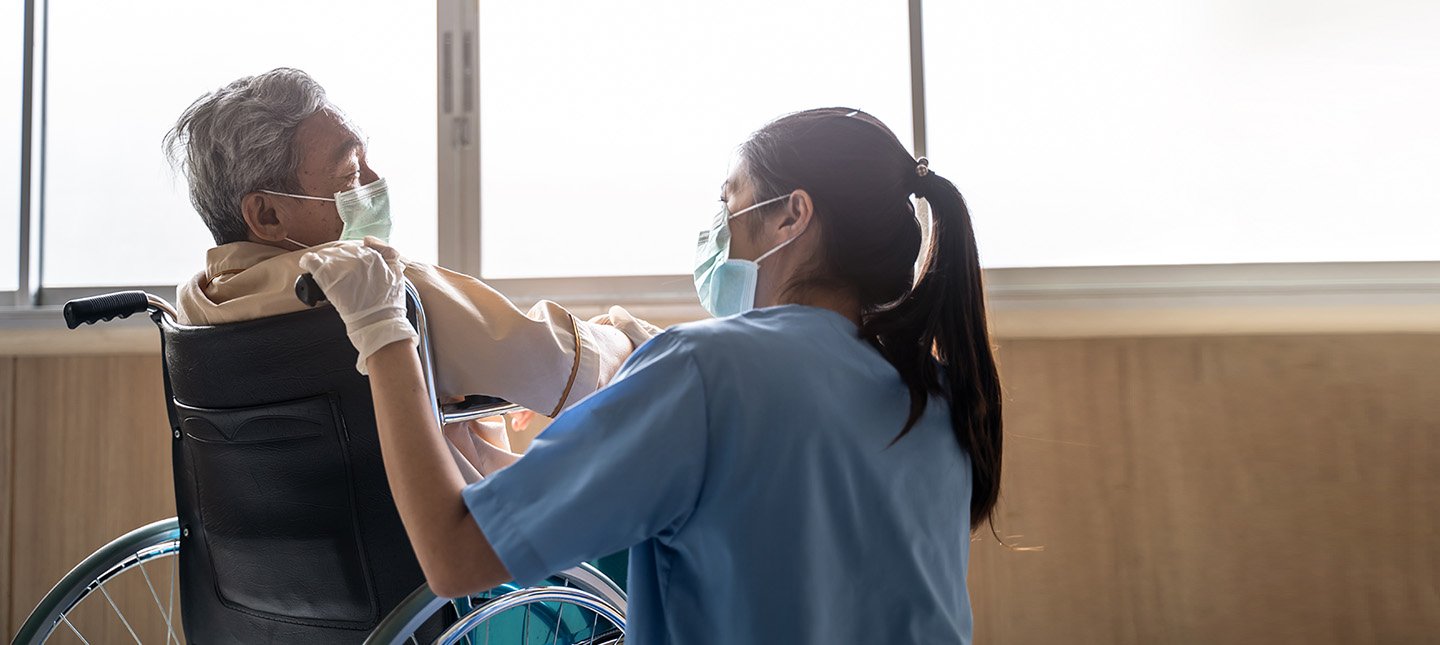 Asian nurse taking care of mature male patient sitting on wheelchair in hospital. Young woman and old man wearing surgical face mask for protection of covid 19 pandemic. Girl smile to elderly man. nursing home ECHO program