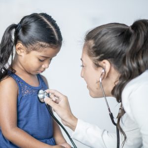 A young hispanic girl looks down at the stethoscope that her doctor is placing on her chest. Her doctor is a young caucasian woman and is wearing professional clothing.