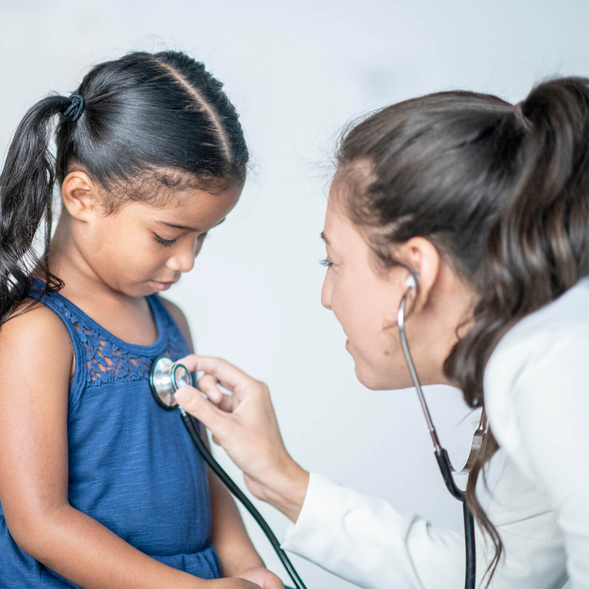 A young hispanic girl looks down at the stethoscope that her doctor is placing on her chest. Her doctor is a young caucasian woman and is wearing professional clothing.
