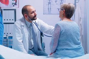 Elderly woman doctor appointment in modern private clinic while she sitts on hospital bed and the physician is listening her heart beat. Health care medicine treatment, specialist consultation